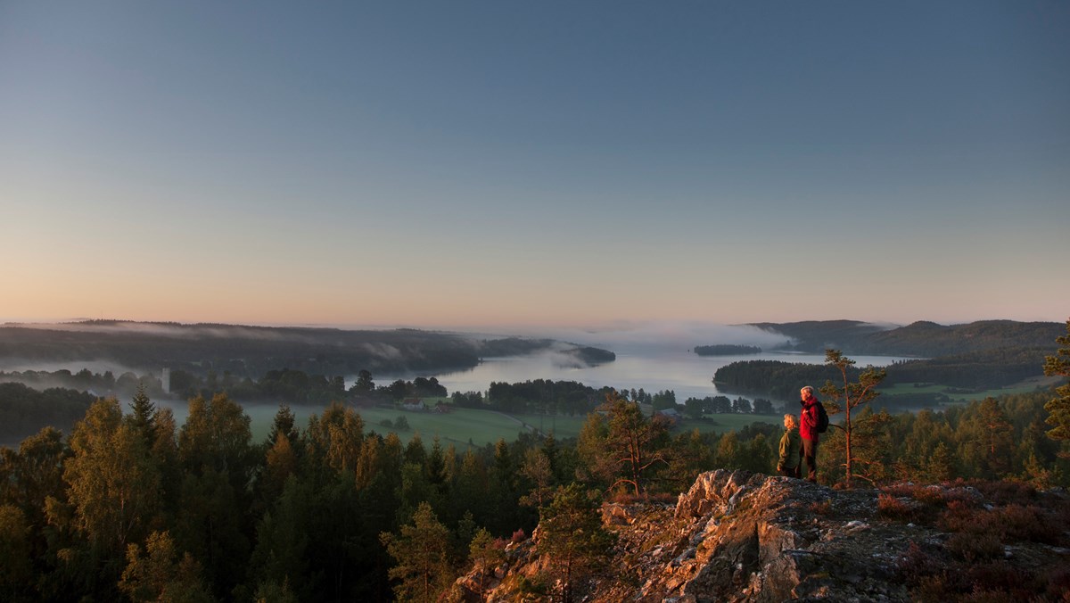 Två personer uppe på en klippa. Utsikt: skog, fält och en sjö med dimma över.