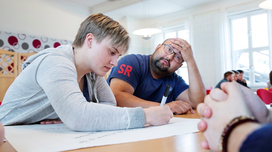 Två folkhögskoledeltagare i ett klassrum. Den ena ser ut att tänka högt och den andra noterar tankarna på ett papper.