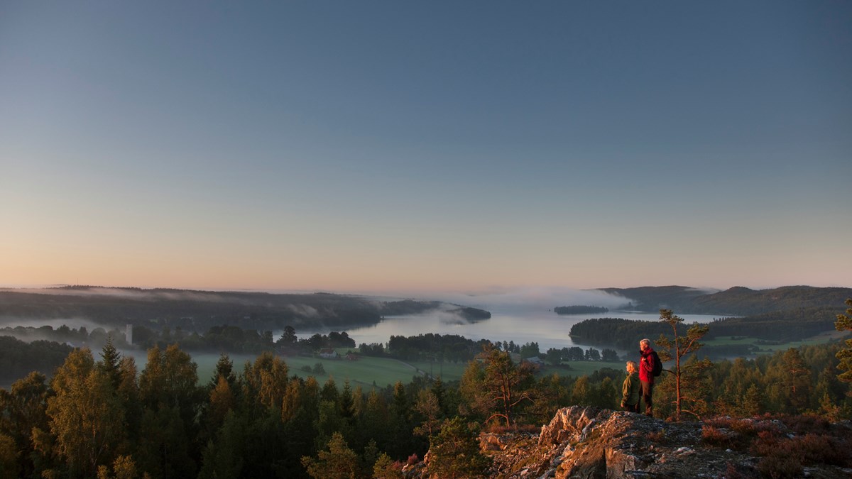 Två vandrare på toppen av en höjd. Utsikten består av skog, fält och en sjö med dimma över.