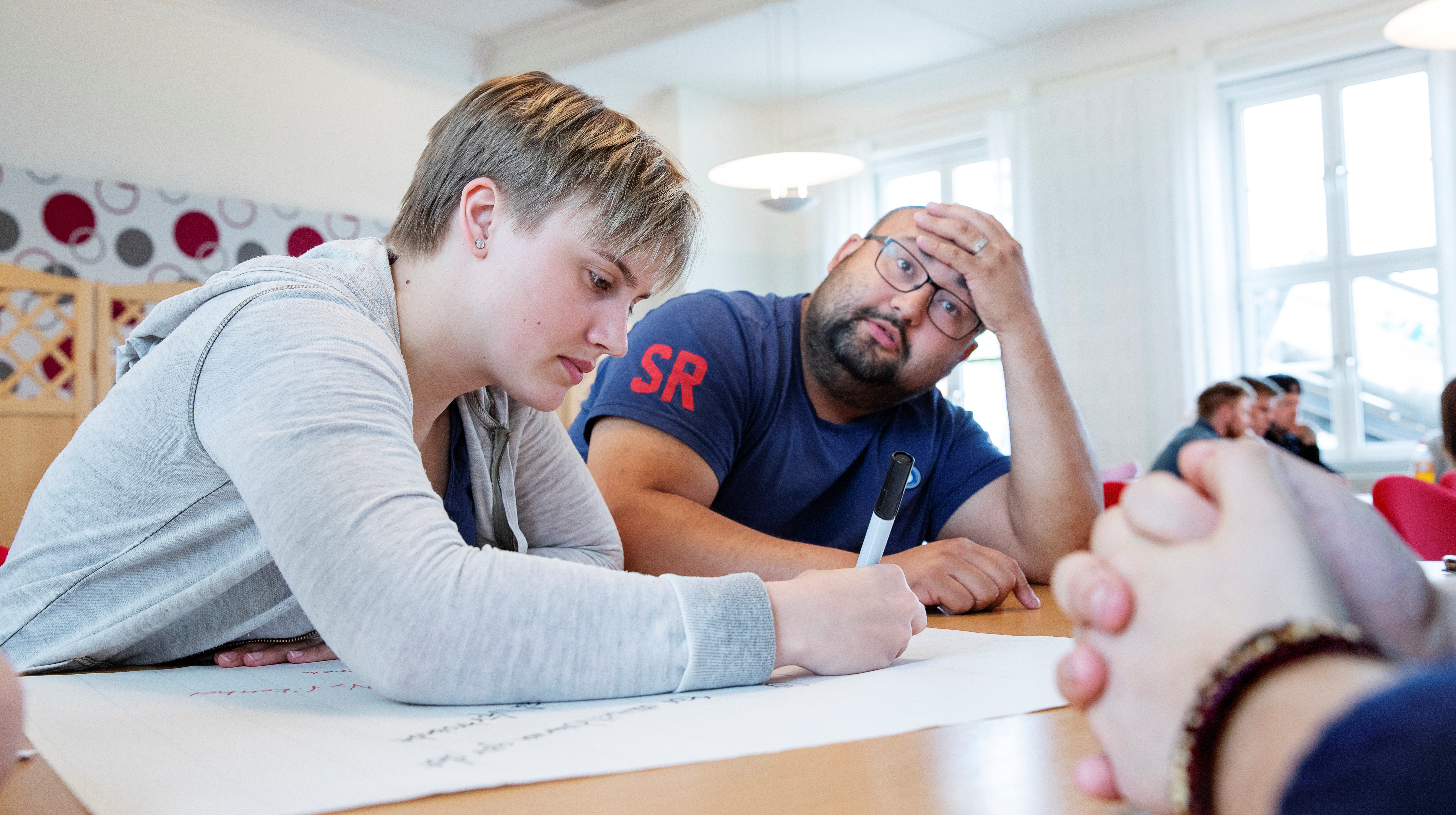 Två folkhögskoledeltagare i ett klassrum. Den ena deltagaren ser ut att fundera högt, medan den andra antecknar.