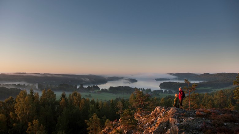 Två vandrare på en klippa ute i Dalslands natur. I bakgrunden syns skog, fält och dimmiga sjöar.