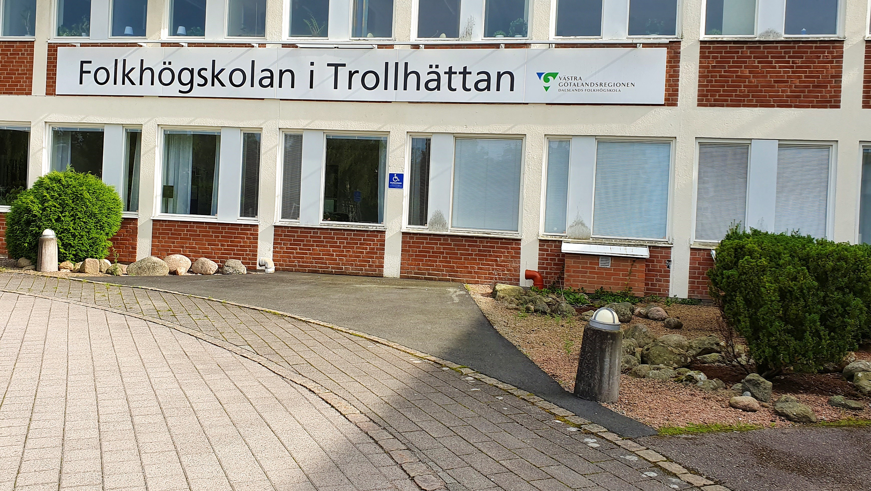 Utsidan av folkhögskolan i Trollhättan. Det är en gammal sjukhusbyggnad i tegel. På huset hänger en skylt där det står Folkhögskolan i Trollhättan.