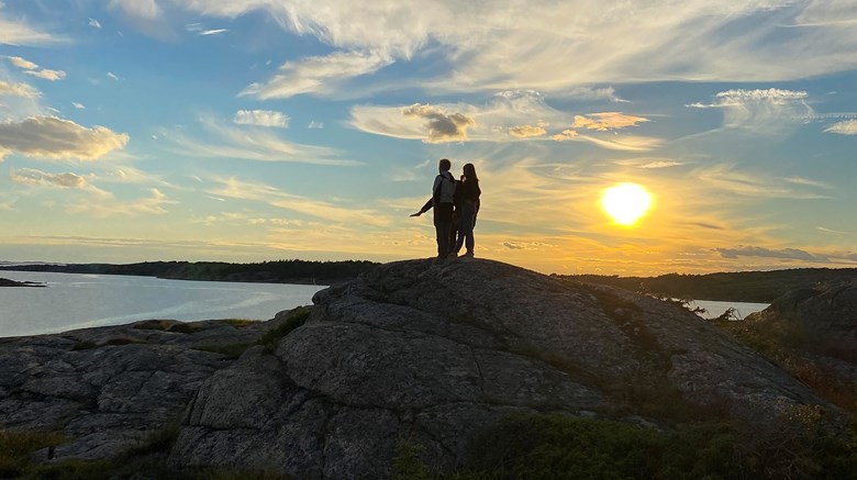Två personer som står på en bergsknalle och tittar ut över havet medan solen går ner.