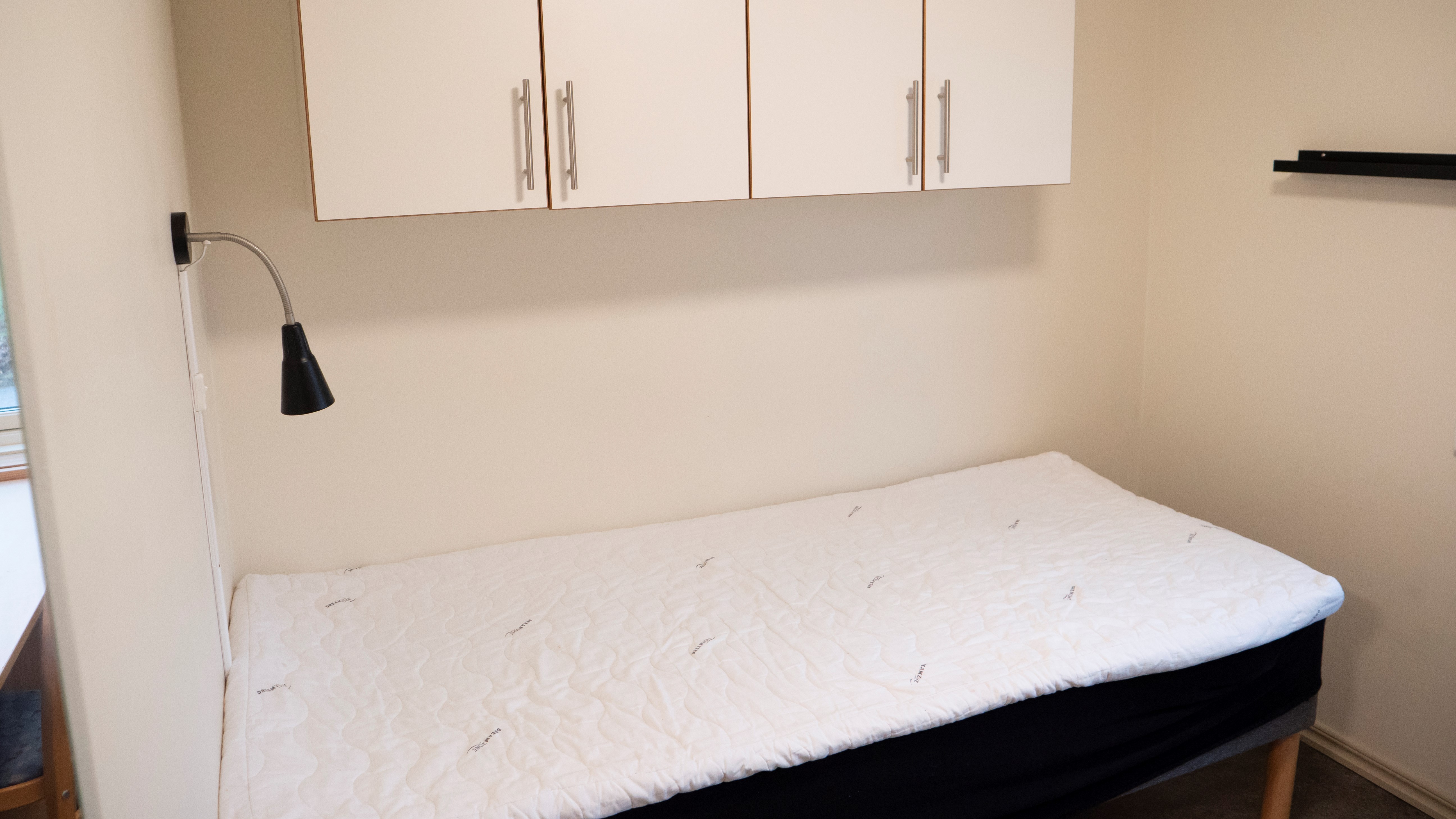 Litet enkelrum, Gråbo, Dalslands folkhögskola. En säng med sänglampa och fyra skåp ovanför.