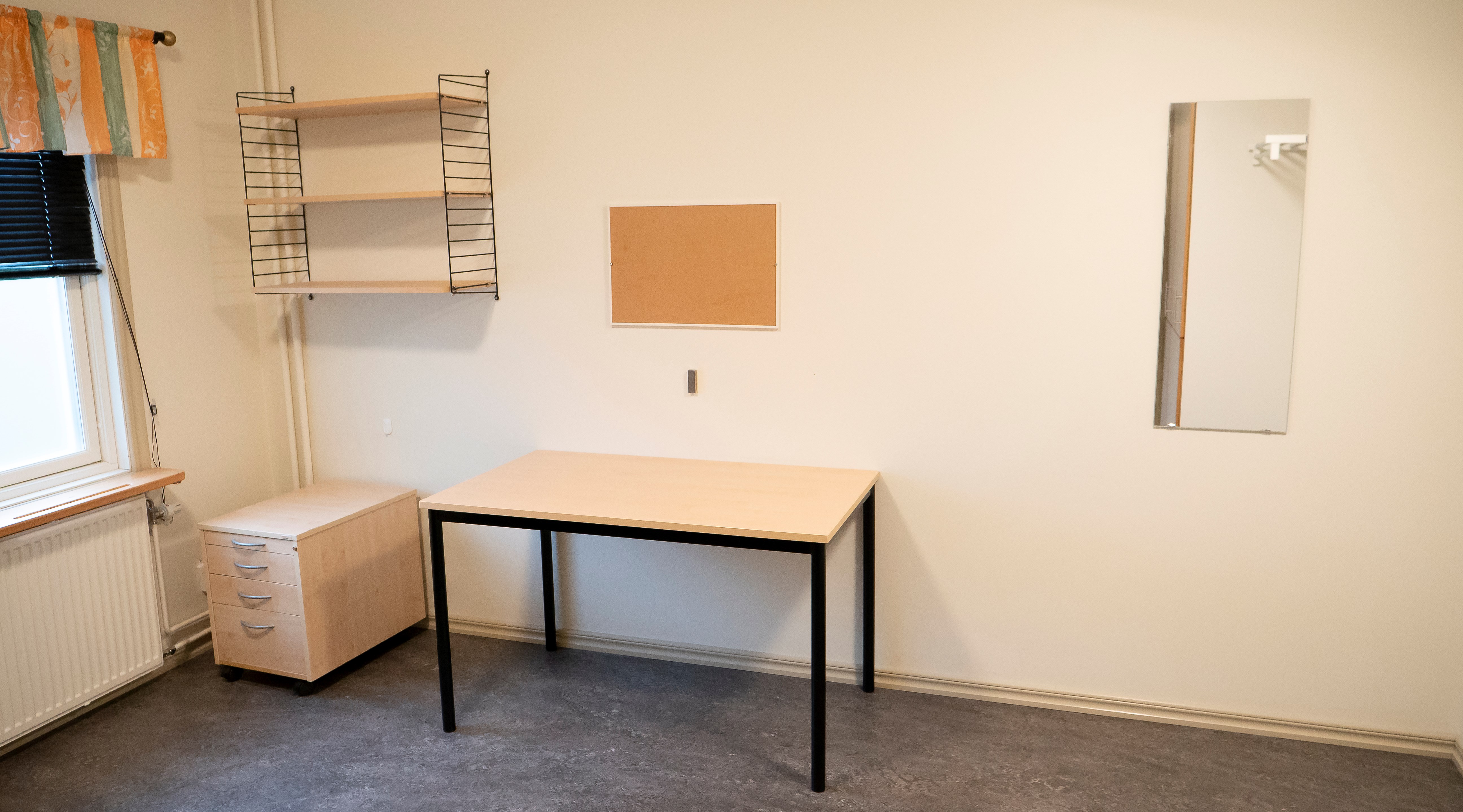 Ett rum med en hurts, skrivbord, hylla, anslagstavla och spegel.