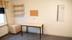 Ett rum med en hurts, skrivbord, hylla, anslagstavla och spegel.