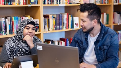 Två folkhögskoledeltagare i ett bibliotek. Den ena sitter vid en dator och den andra vid en surfplatta.