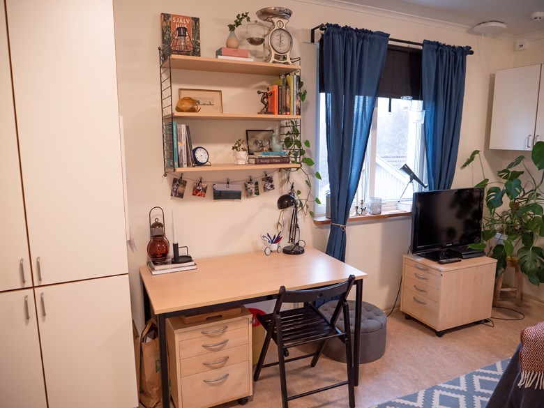 Ett skrivbord med en hylla ovanför, samt en tv på en hurts.